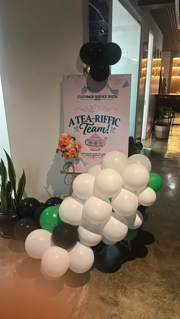 Balloon garlands in black, white and green framing a Customer Service Week 2026 A Tea-riffic Team poster at an office reception corner