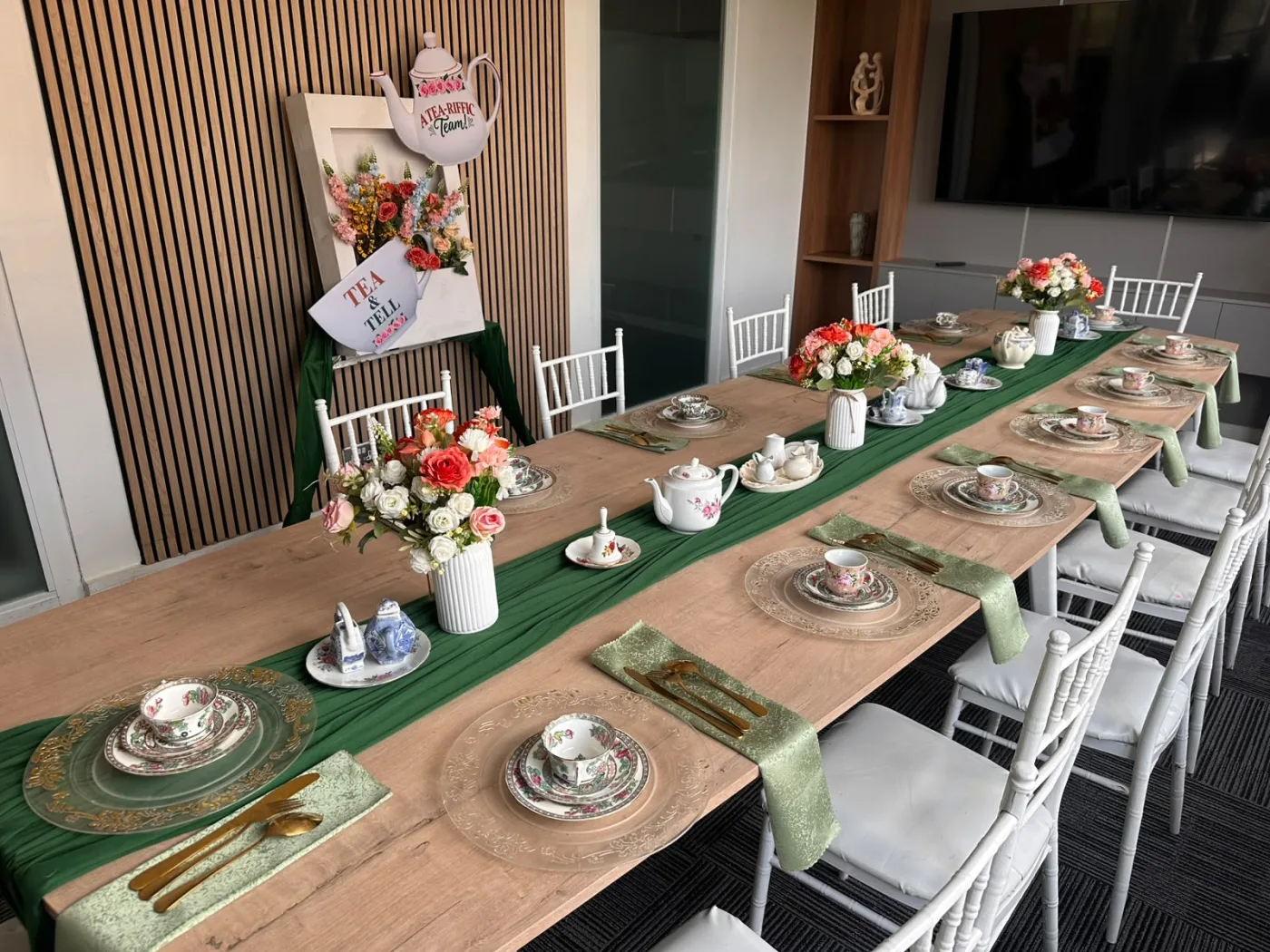Long boardroom table dressed for a high tea with white chiavari chairs, a green silk runner, vintage floral china place settings and fresh pink and white floral arrangements down its length