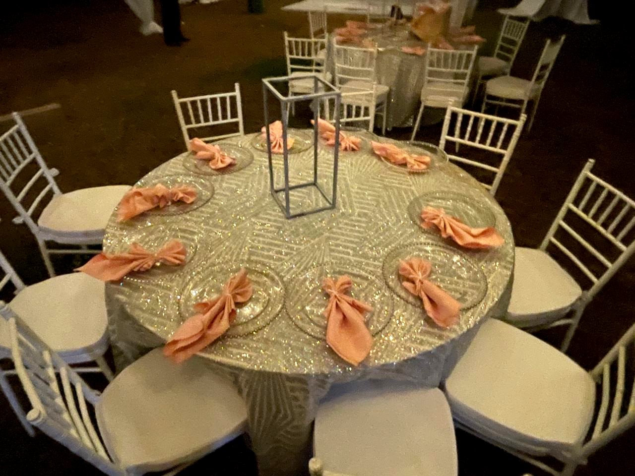 Guest round dressed in patterned silver sequin linen with glass charger plates, peach napkins tied at each setting, a tall geometric centrepiece, and white chiavari chairs