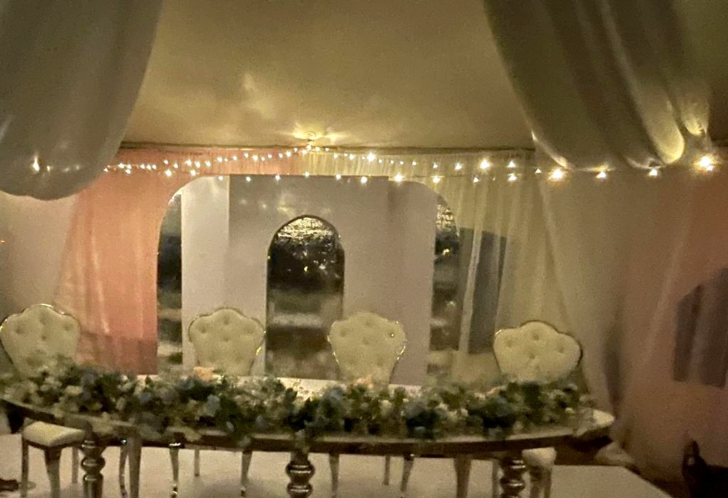 Head table at an evening marquee reception, silver mirrored table with a runner of white and dusty blue florals, four ivory tufted high-back chairs, a white and mirrored arch backdrop, blush drapes and fairy lights overhead