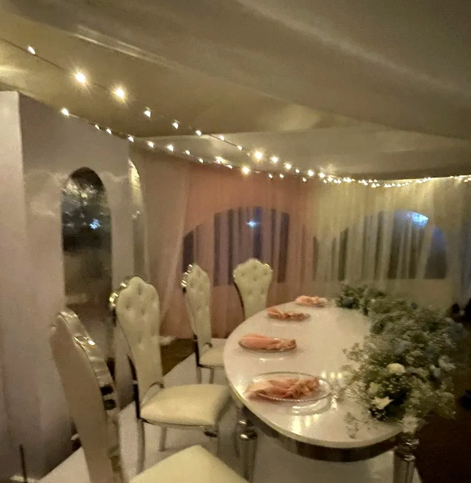 Three-quarter view along the mirrored head table, ivory tufted chairs, white and dusty blue floral runner, peach napkins on glass charger plates, blush drapes and fairy lights behind