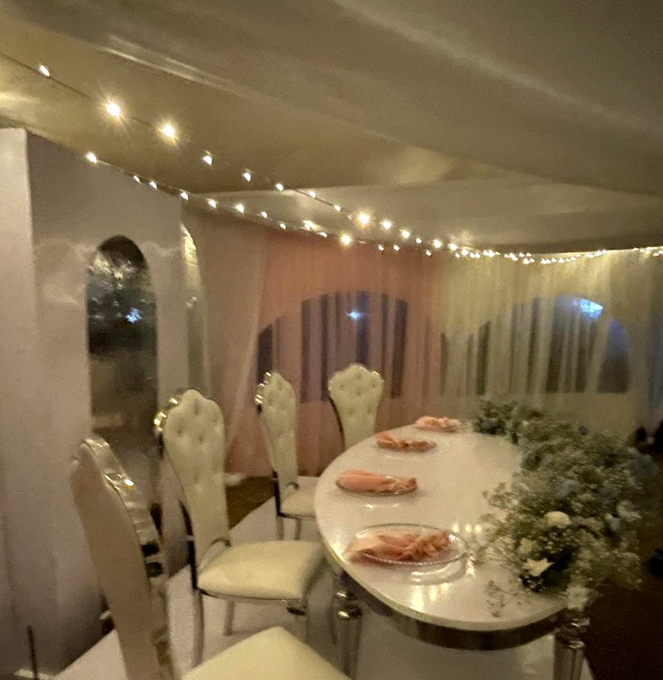 Three-quarter view along the mirrored head table, ivory tufted chairs, white and dusty blue floral runner, peach napkins on glass charger plates, blush drapes and fairy lights behind