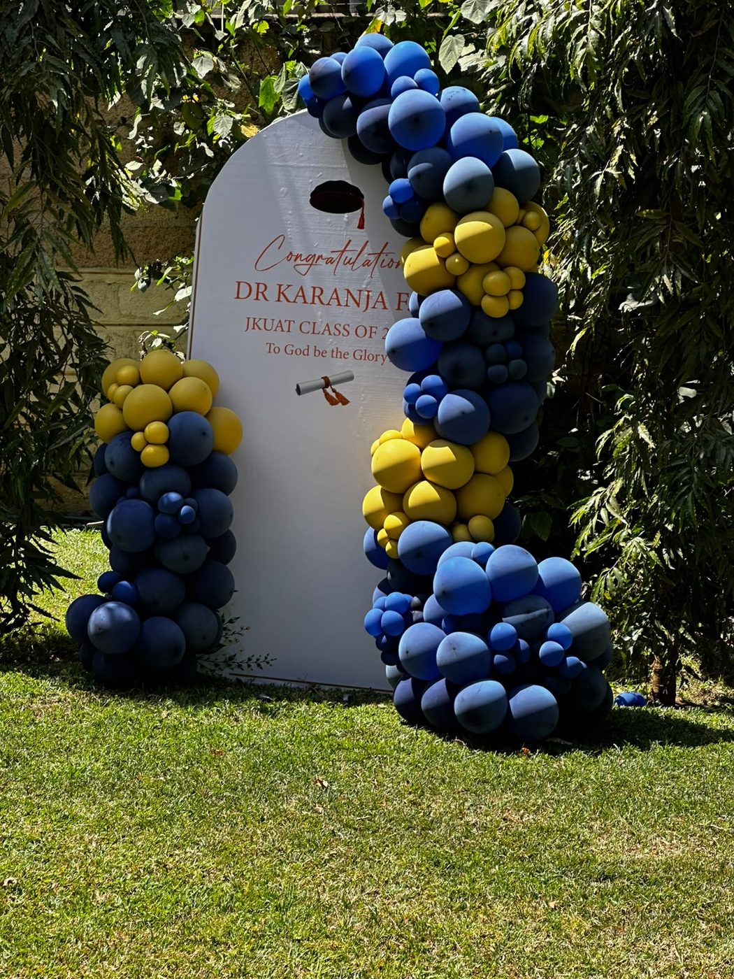 Organic balloon arch in navy, royal blue and yellow framing a white arched backdrop that reads Congratulations Dr Karanja, JKUAT Class of, To God be the Glory, with a graduation cap and scroll motif, set on clipped lawn against greenery