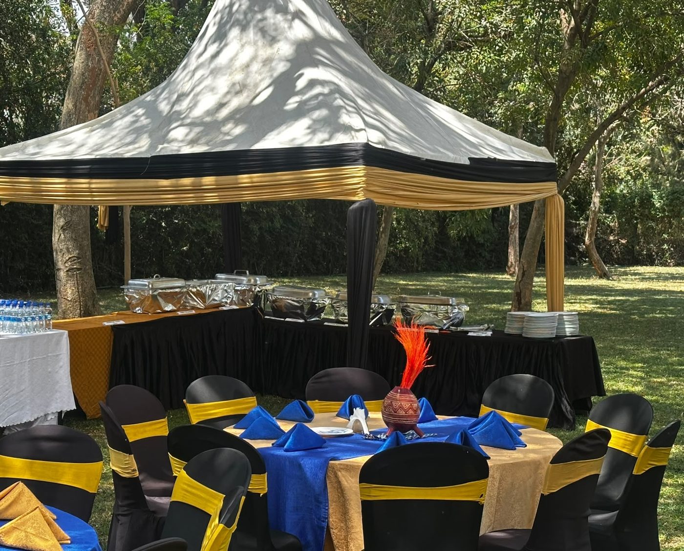 Buffet gazebo draped in black and gold with chafing dishes, stacked plates and a drinks table of bottled water and sodas, guest tables in blue and gold in the foreground