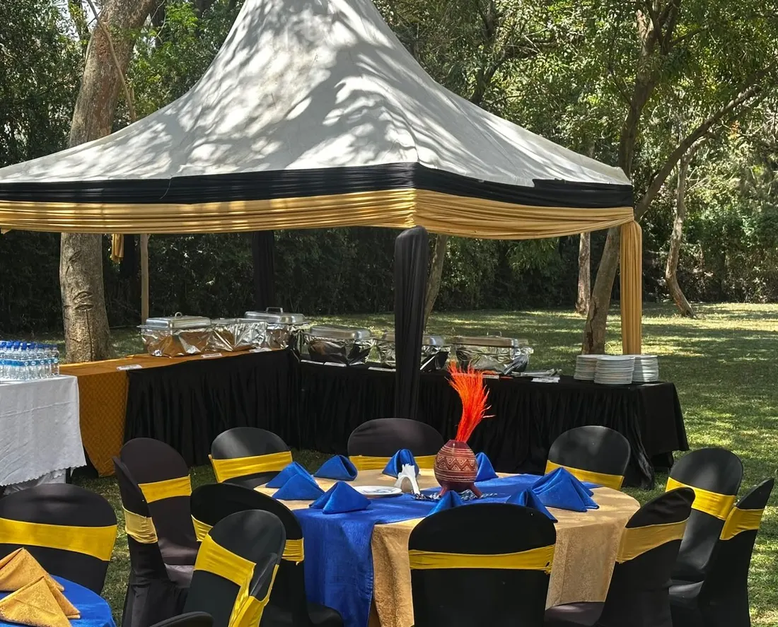 Buffet gazebo draped in black and gold with chafing dishes, stacked plates and a drinks table of bottled water and sodas, guest tables in blue and gold in the foreground