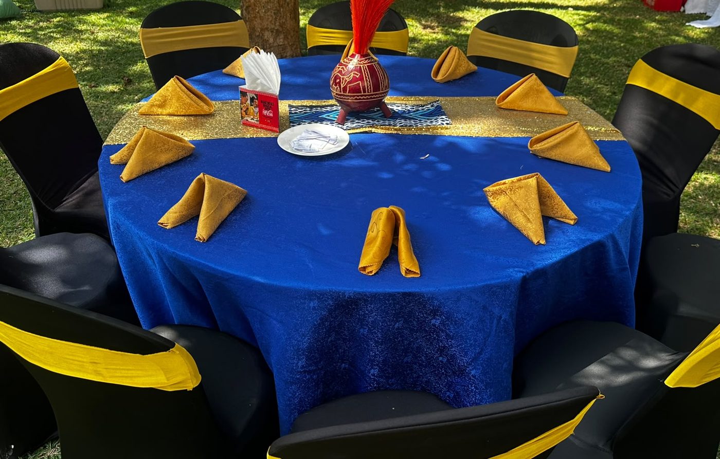 Overhead view of a single guest round in royal blue velvet with a gold sequin runner, gold napkins folded at each setting, and a decorated terracotta gourd with a tall red feather plume on a printed kanga runner