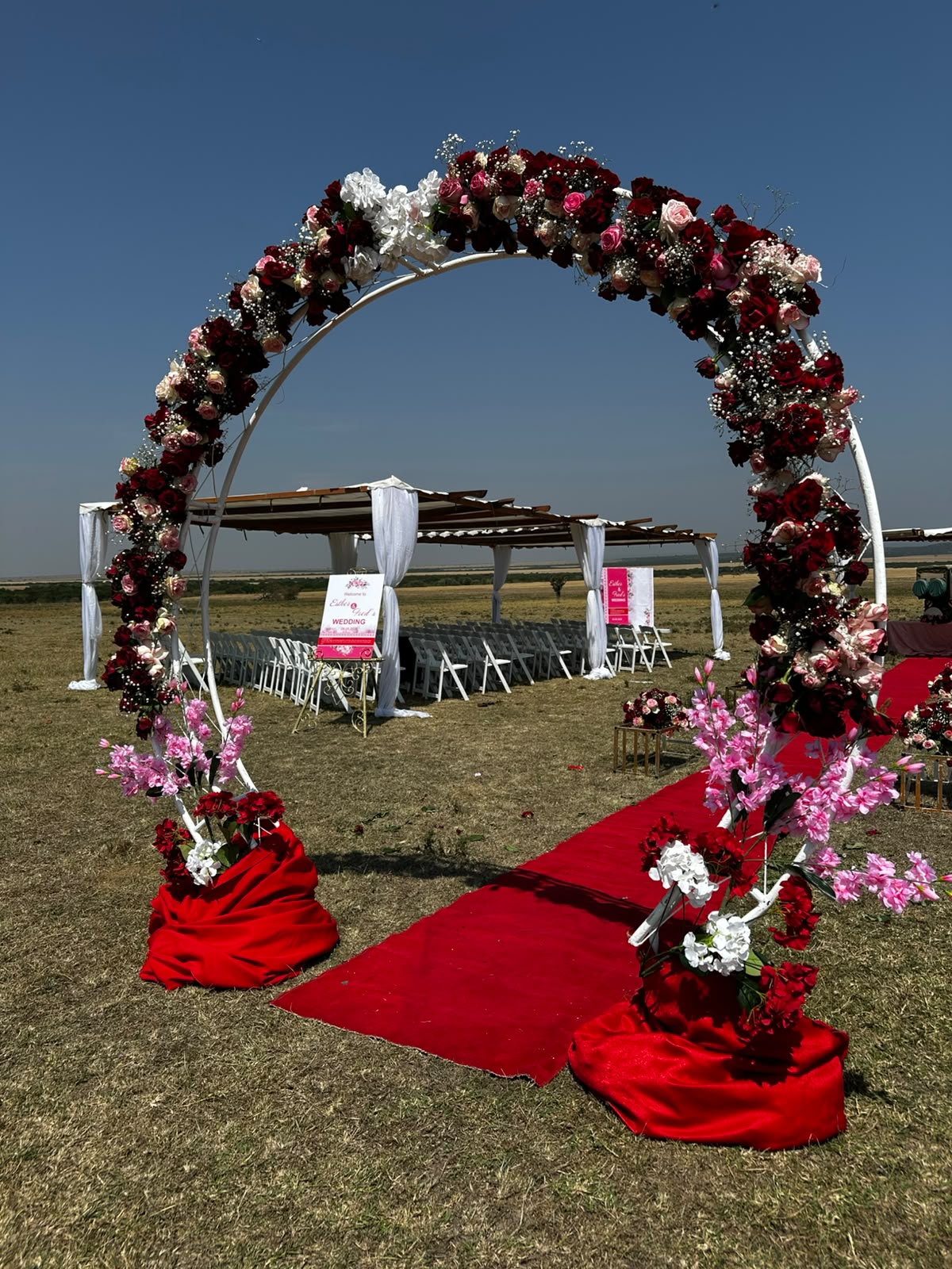 Circular floral moon arch covered in deep red and blush roses with baby's breath, standing over a red carpet aisle on open Maasai Mara grassland, ceremony chairs and a draped pergola in the distance