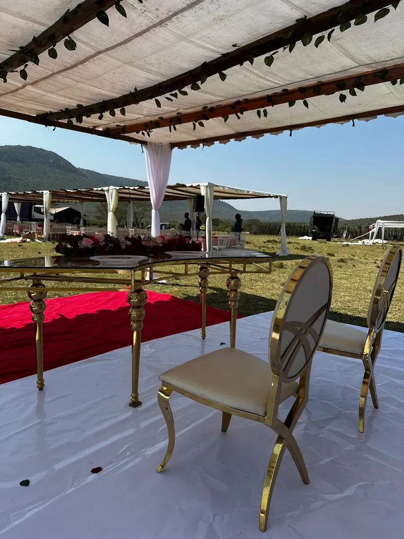 Gold oval-back chairs with cream upholstery at a gold-framed glass dining table under a canvas pergola trimmed with green garland, escarpment hills on the horizon behind