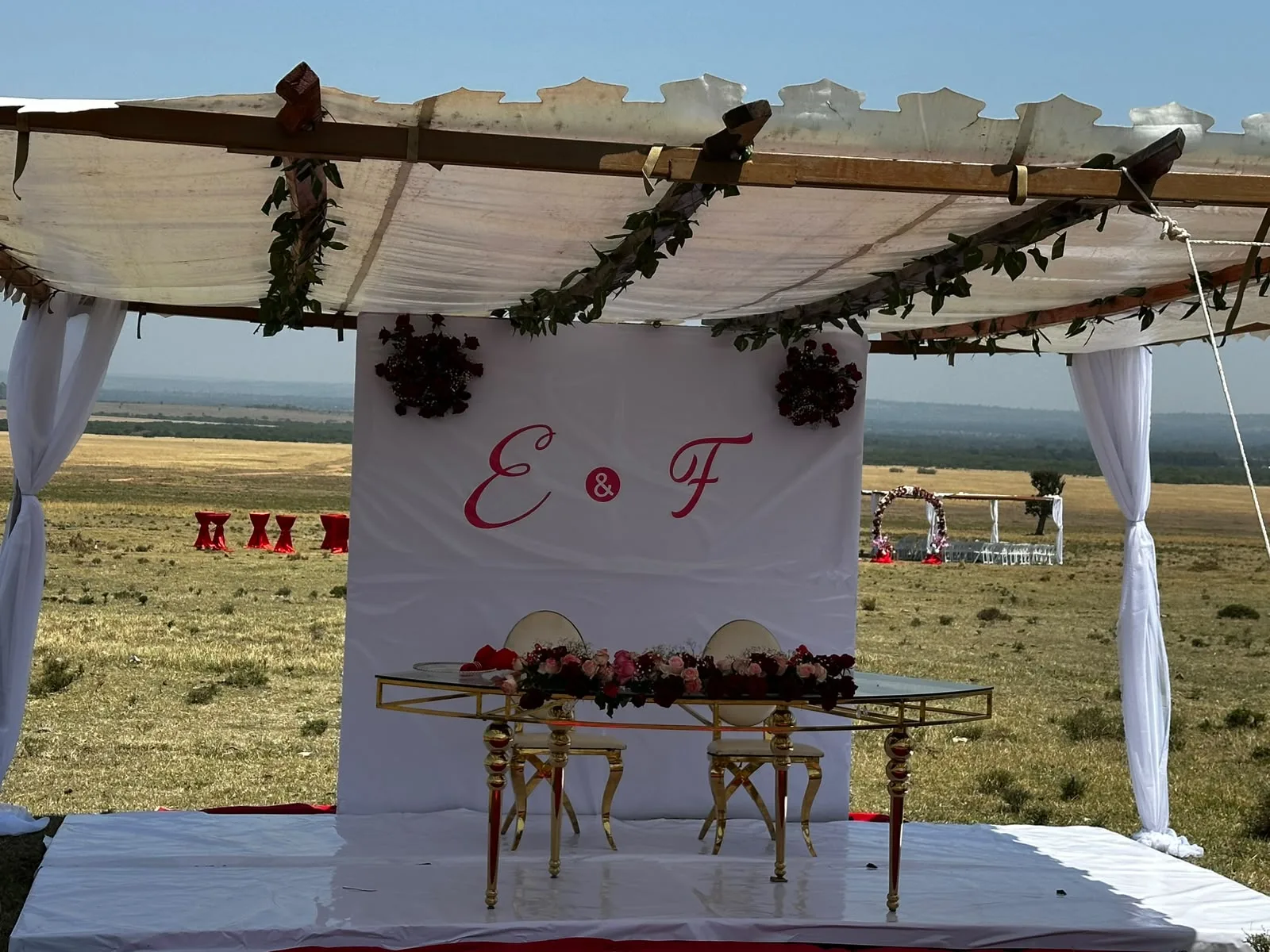 Draped ceremony pergola on a raised platform with the floral arch and guest seating far off across the Maasai Mara plain
