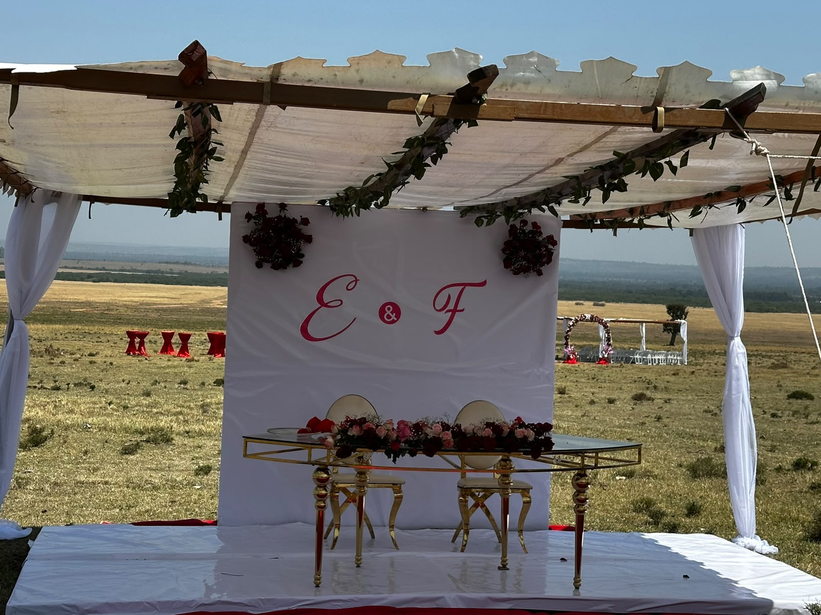 The ceremony pergola with white draping and green garland, an E and F monogram backdrop and a gold sweetheart table on a raised white platform, with the floral arch and rows of ceremony chairs small in the distance across the Maasai Mara plain