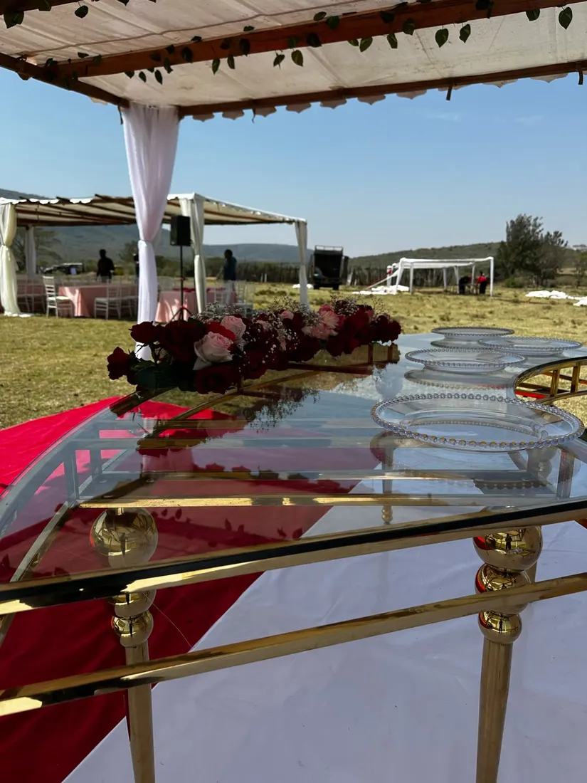 Close view along a glass dining table showing beaded charger plates and a low runner of deep red and blush roses with baby's breath, red carpet and further pergolas beyond