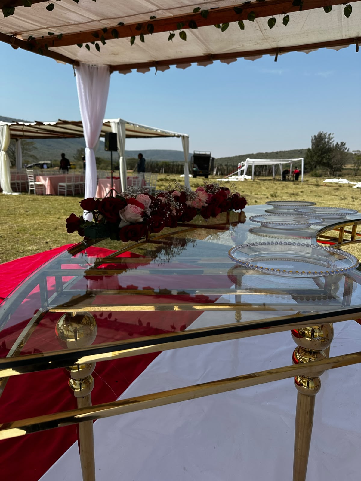 Close view along a glass dining table showing beaded charger plates and a low runner of deep red and blush roses with baby's breath, red carpet and further pergolas beyond