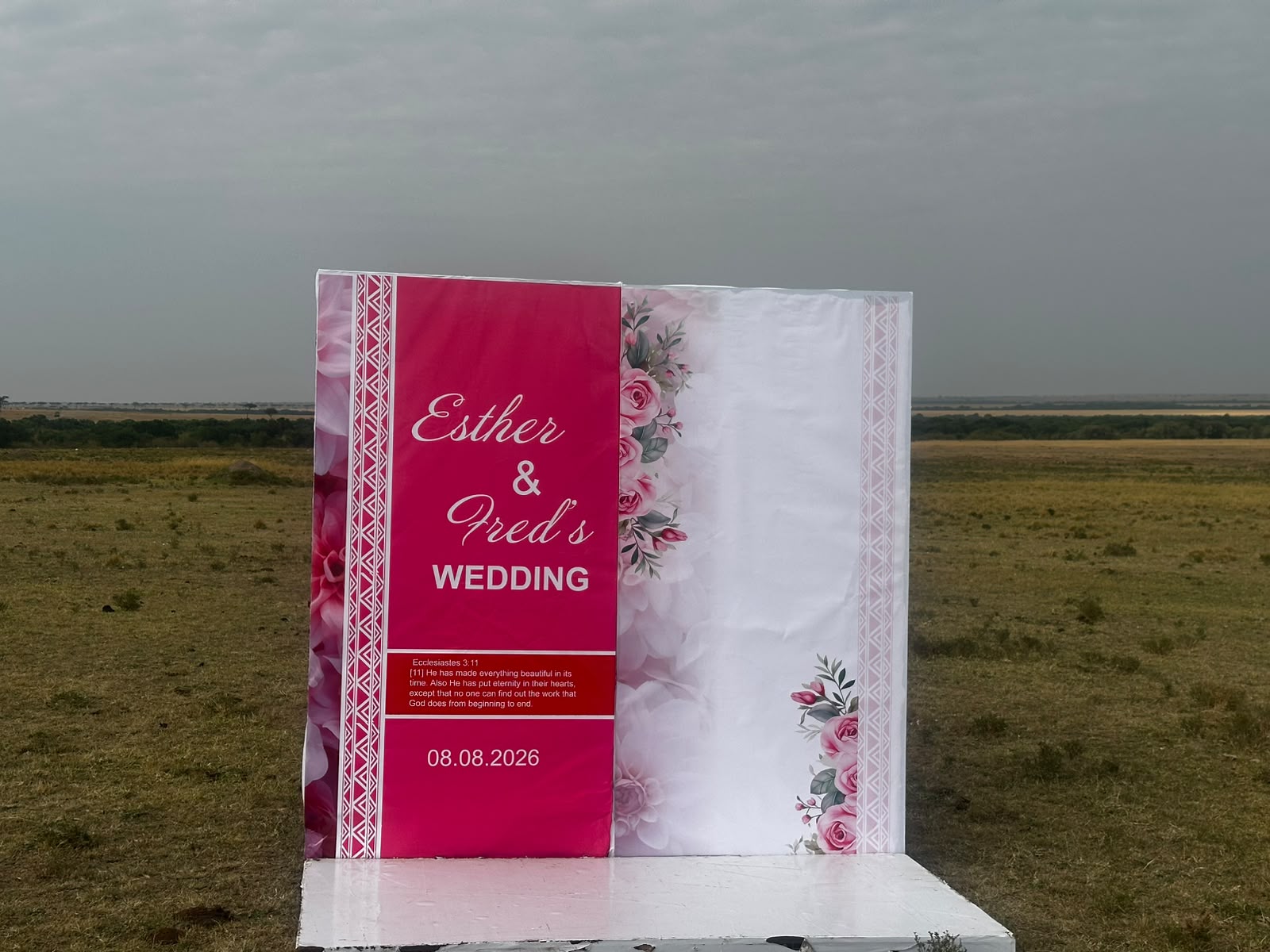 Freestanding welcome backdrop in crimson and blush florals reading Esther and Fred's Wedding with Ecclesiastes 3:11 and the date 08.08.2026, standing alone on open grassland under an overcast sky
